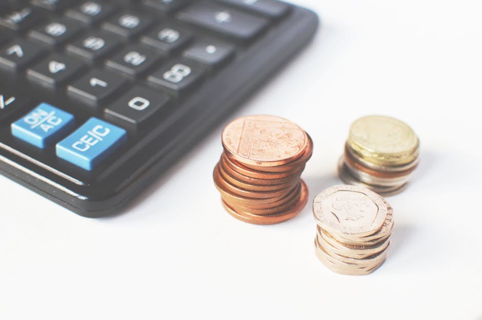 Close-up image showing a black calculator with blue and white keys, positioned in the upper left corner of the frame. To the right of the calculator, there are three distinct stacks of euro coins arra