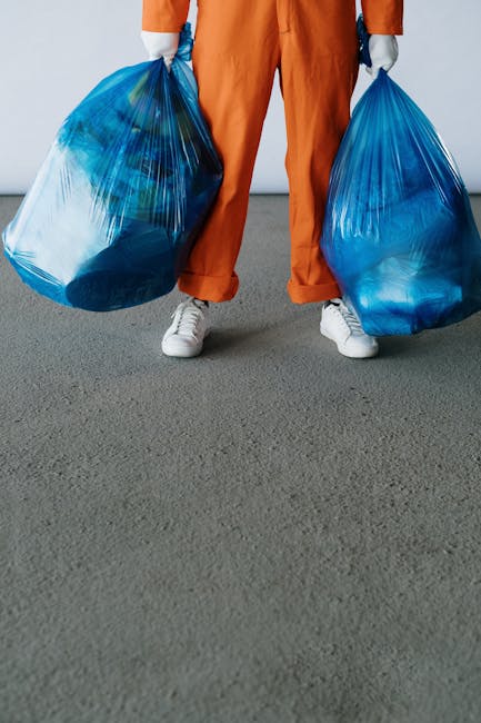 A person dressed in orange work pants and white sneakers standing on a dark grey outdoor surface, holding two large blue plastic rubbish bags filled with waste materials. The bags are semi-transparent